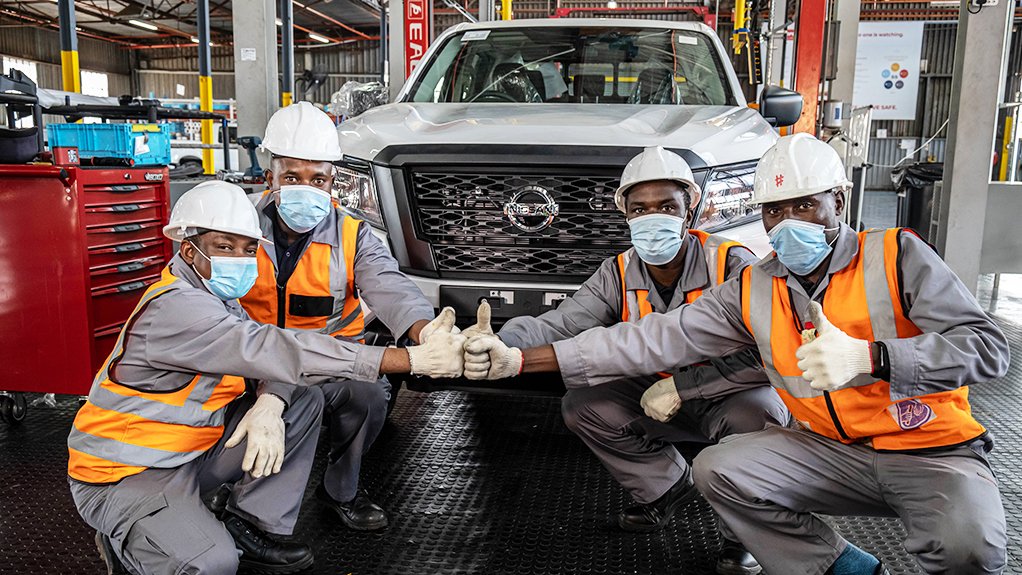 Four factory workers in hard hats and masks give thumbs up in a vehicle assembly bay in front of a Nissan pickup truck.
