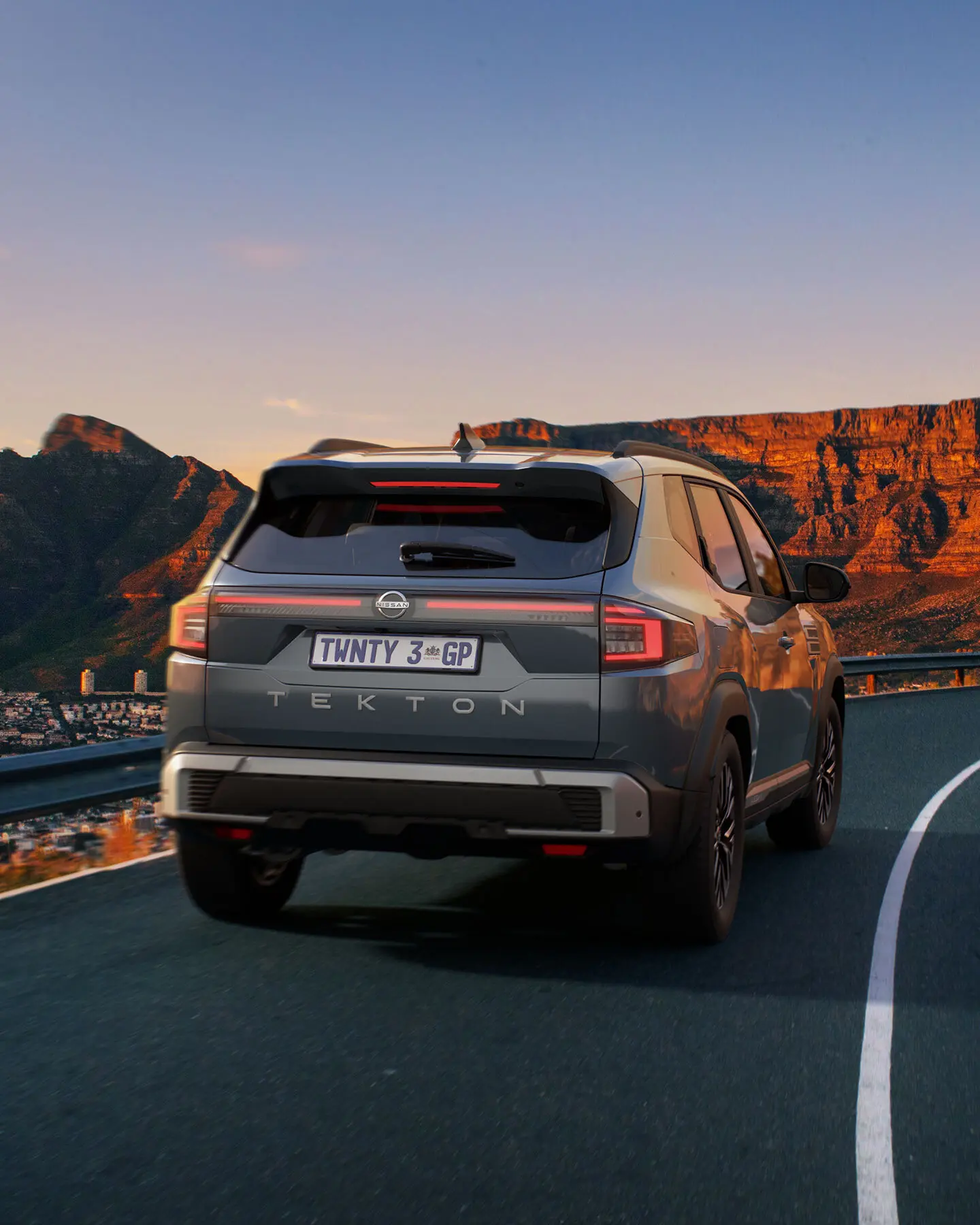 Rear view of a silver Nissan Tekton SUV on a winding mountain highway at sunset, with orange rock cliffs in the background and a visible license plate TWNTY 3 GP.