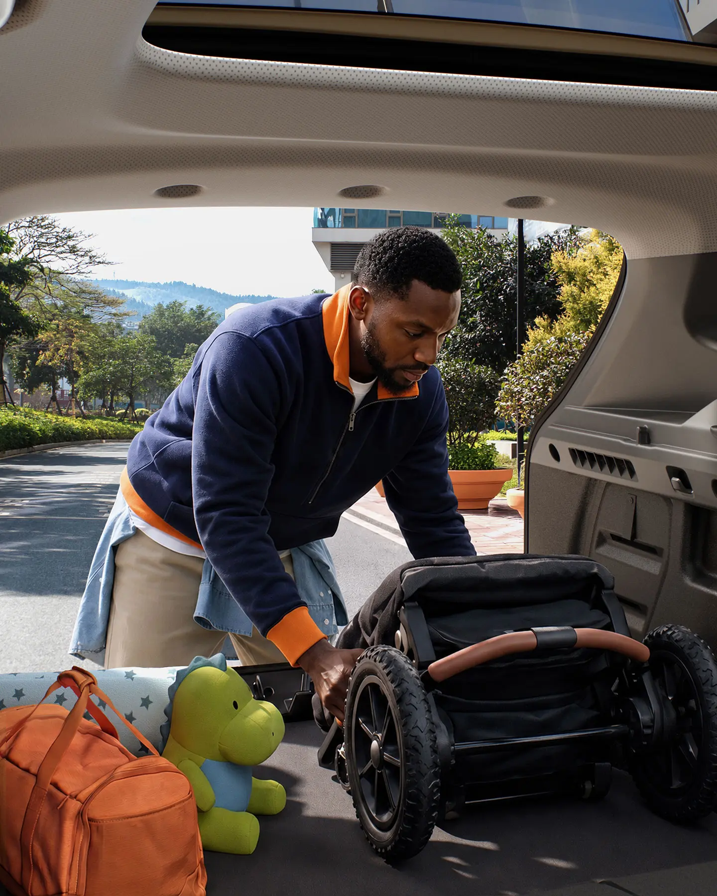 Man loading a baby stroller into the open trunk of an SUV in a parking lot, with an orange backpack and a green turtle plush nearby.